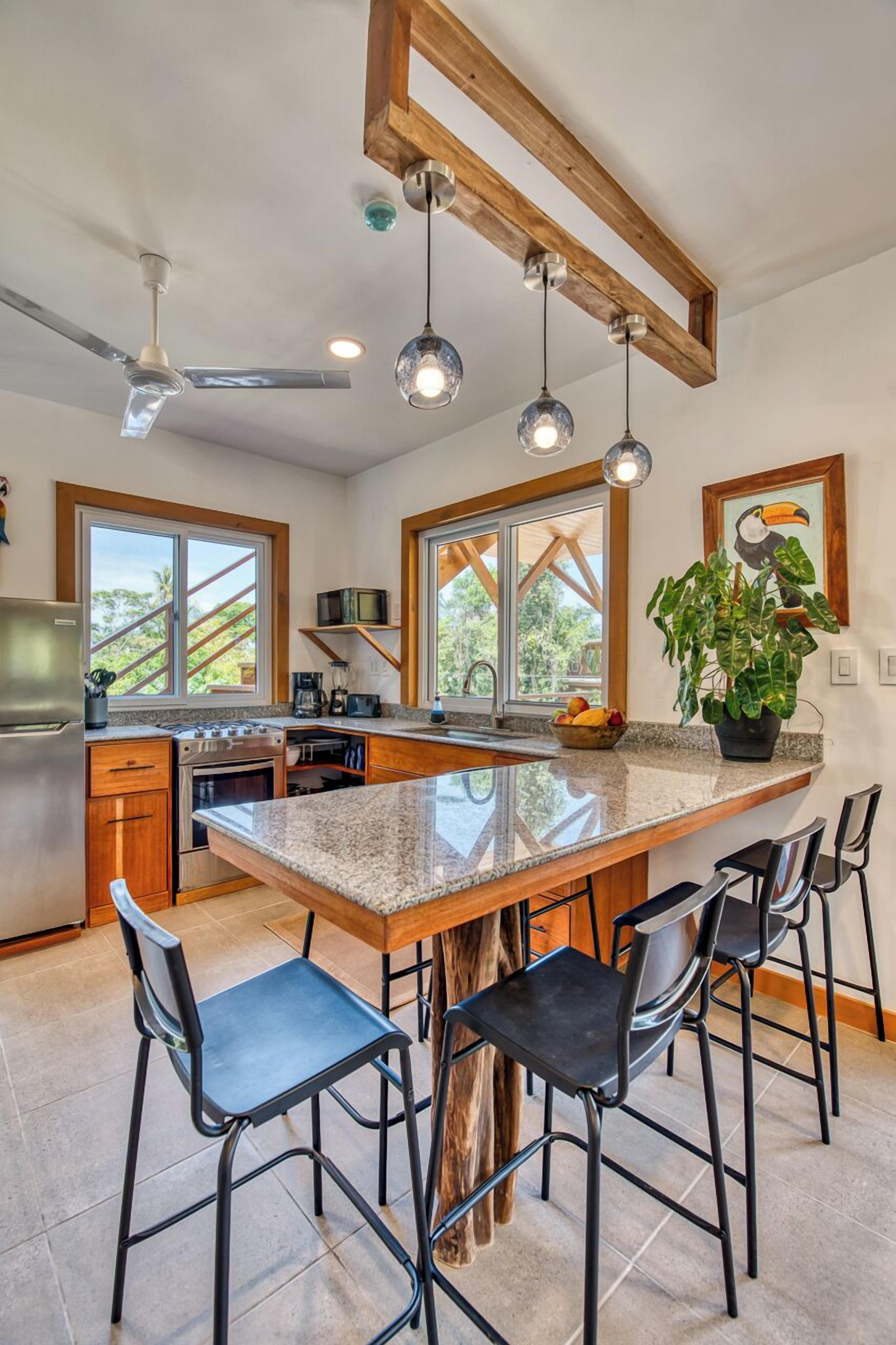 A modern kitchen with a large island, bar stools, a ceiling fan, pendant lights, and a view of the outdoors.