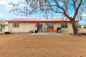 A single-story building with a red roof, a covered patio area with two orange chairs, and a large tree in front.