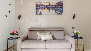 Ein beiges Sofa mit einem gemusterten Kissen, zwei Beistelltische mit Topfpflanzen und ein an der Wand hängendes Bild von einer Brücke.