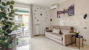 Ein Wohnzimmer mit beiger Couch, einer Uhr, einer Wand-Klimaanlage, einem kleinen Tisch mit Blumenvase und Blick auf ein Gebäude durch eine Glastür.