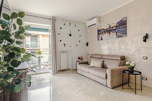 A living room with a beige sofa, a clock, a wall-mounted air conditioner, a small table with a flower vase, and a view of a building through a glass door. - [luggage storage] - La Bellavita Trastevere (Roma)