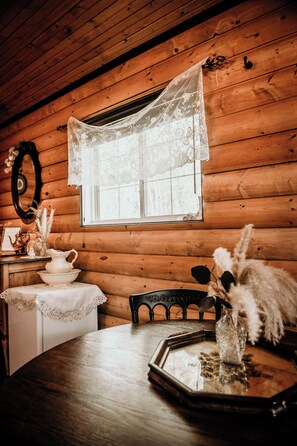 A wooden cabin interior with a round table, lace window treatment, and a mirror on the wall.