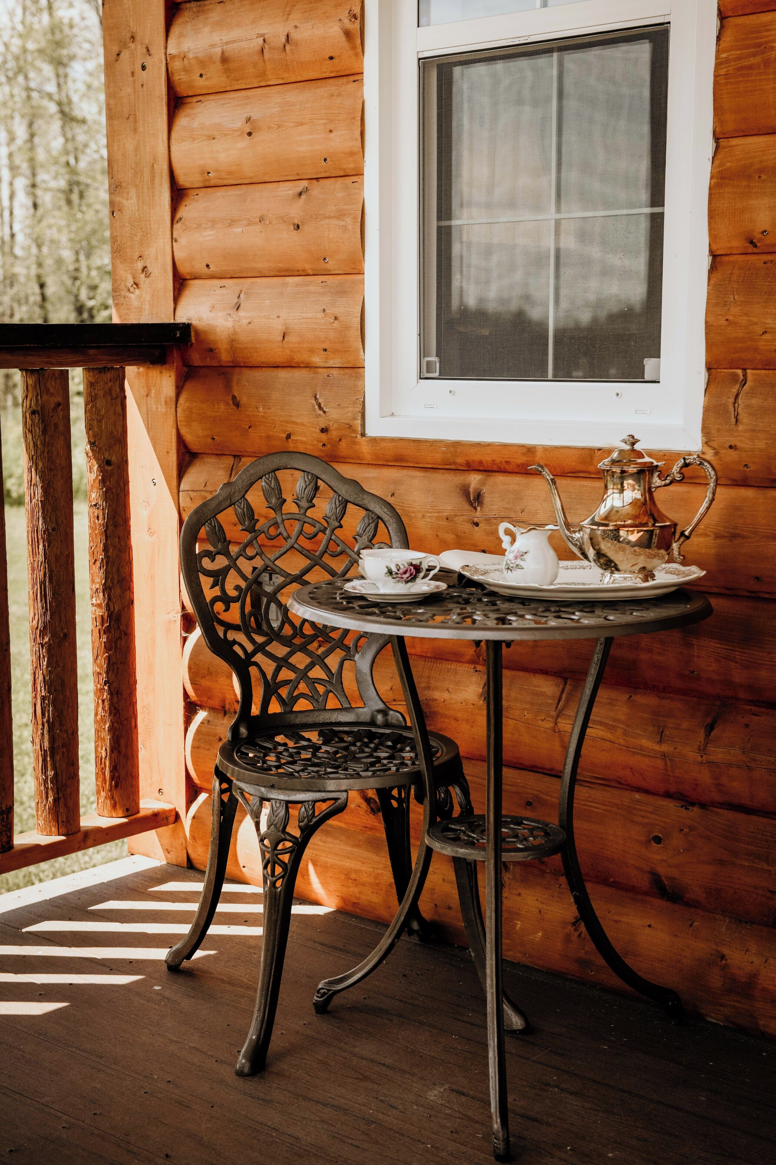A wooden cabin with a window, a metal table with a chair, and a tea set on the table.
