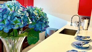 Une cuisine avec des meubles de couleur rouge, un plan de travail blanc, un vase rempli de fleurs bleues et une rangée d’assiettes accompagnées de serviettes bleues.