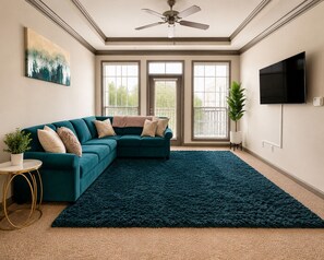 A living room with a sectional sofa, a ceiling fan, a large window with blinds, a flat-screen TV, and a potted plant.