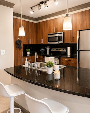 A modern kitchen with wooden cabinets, stainless steel appliances, a black granite countertop, and pendant lighting.