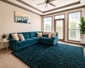 A teal sectional sofa, a dark blue rug, a ceiling fan, and large windows with blinds.