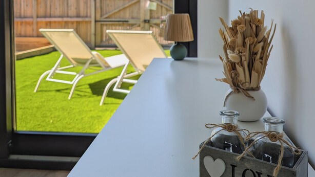 Une pièce donnant sur un jardin, avec une table blanche, une lampe et un vase décoratif contenant des plantes séchées.