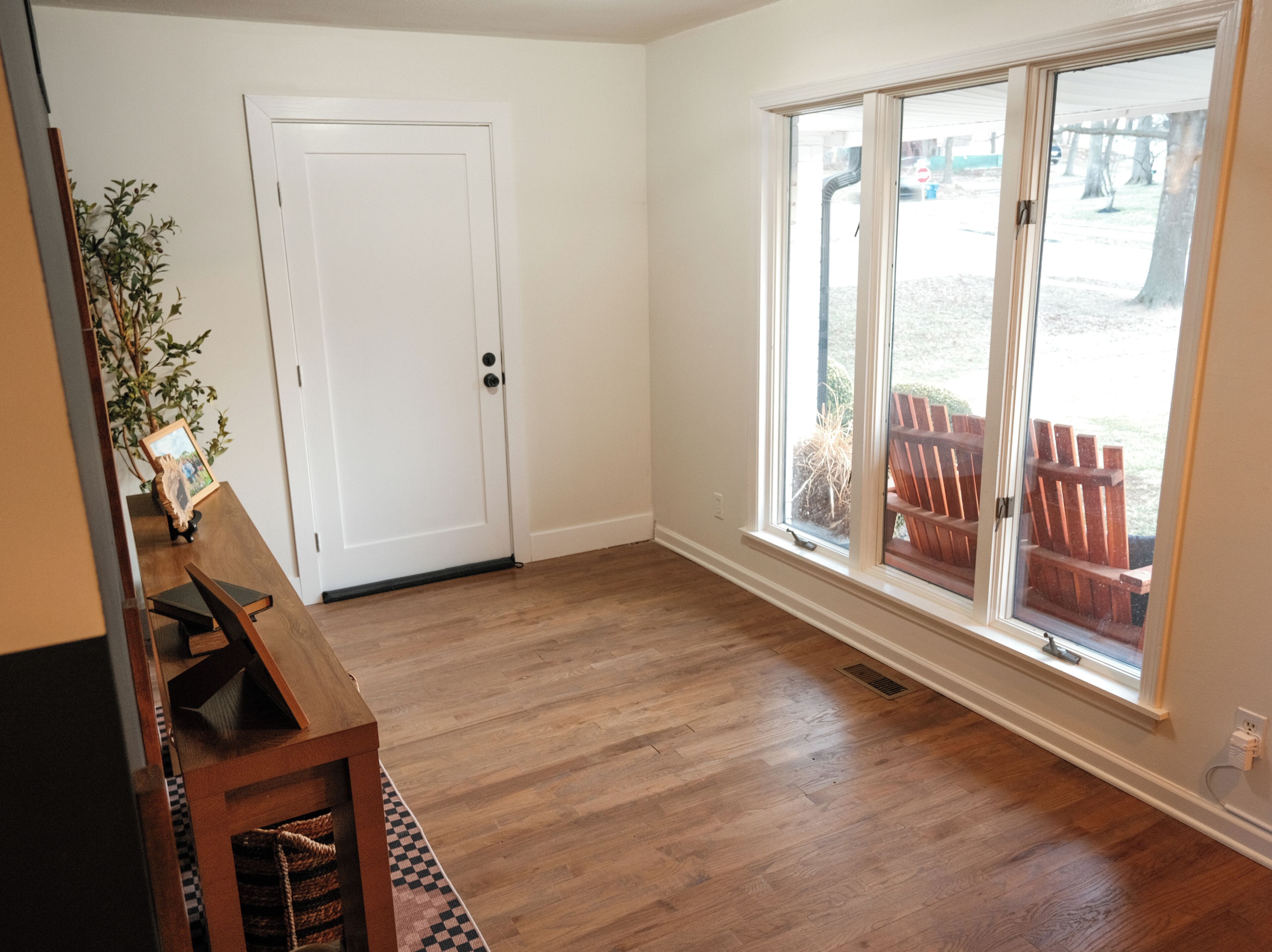 A room with hardwood flooring, a white door, a window with a view of outdoor furniture, and a small table with a plant.