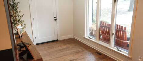 A room with hardwood flooring, a white door, a window with a view of outdoor furniture, and a small table with a plant.