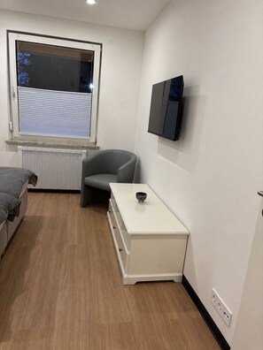 A modern room with a bed, a chair, a white bedside table, a radiator, a window, and a mounted television. - Renovated vacation apartment for up to 5 persons (Bodenwerder)