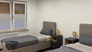 Moderni hotellihuone, jossa on kaksi erillistä sänkyä, pieni yöpöytä lampun kanssa, ikkuna, jossa on sälekaihtimet, ja patteri.