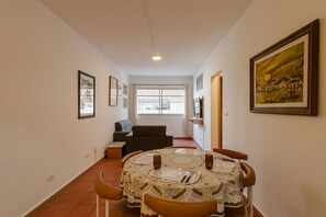 A dining area with a table set for four, a sofa, and a window with curtains. - Apto p 6 Pessoas a 400m da Praia das Pitangueiras (Guarujá)