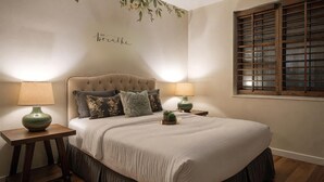 Ein Schlafzimmer mit einem gesteppten Kopfteil, einer Blumen-Wanddekoration, zwei Nachttischen mit Lampen, einem Fenster mit Holzjalousien und einem Bett mit beigefarbener Bettdecke und dunkelbraunen Beinen.