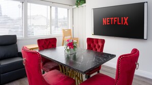 Een moderne kamer met een zwarte tafel, rode stoelen, een prikbord met 'NETFLIX' erop, een zwarte bank en een raam met zonwering.