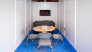 Een kamer met een blauwe vloer, een ronde tafel met stoelen, een bank en een wandgemonteerde televisie waarop 'NETFLIX' wordt weergegeven.