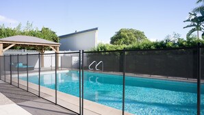 Una piscina con valla de seguridad, una zona de estar cubierta y un edificio al fondo.