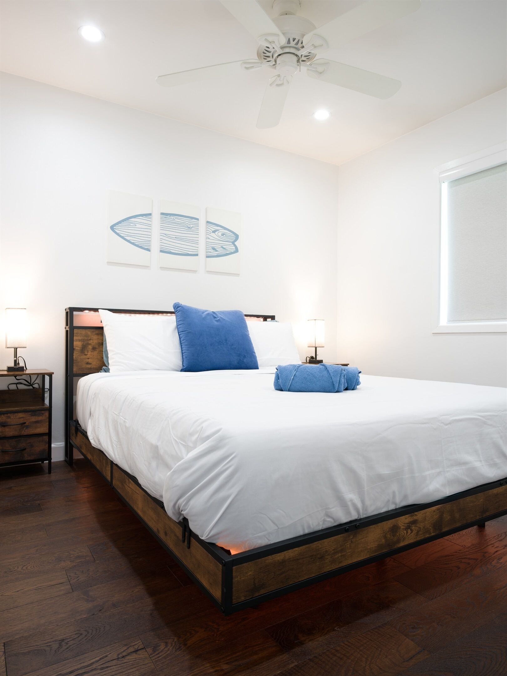 A bedroom with a bed, wooden headboard, white bedding, a ceiling fan, and artwork on the wall.