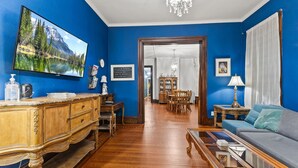A room with a wooden floor, a blue wall, a television, a sofa, a chandelier, and a dining area in the background. - The Duchess Guest House, Walk to Downtown Anderson, Historic! (Anderson)