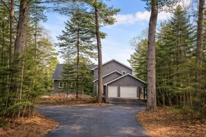 A two-story house with a garage, surrounded by tall trees and a driveway. - Lake Huron Beach House 10 minutes from Mackinaw City, Kayaks, Gym, Amazing Views (Cheboygan)
