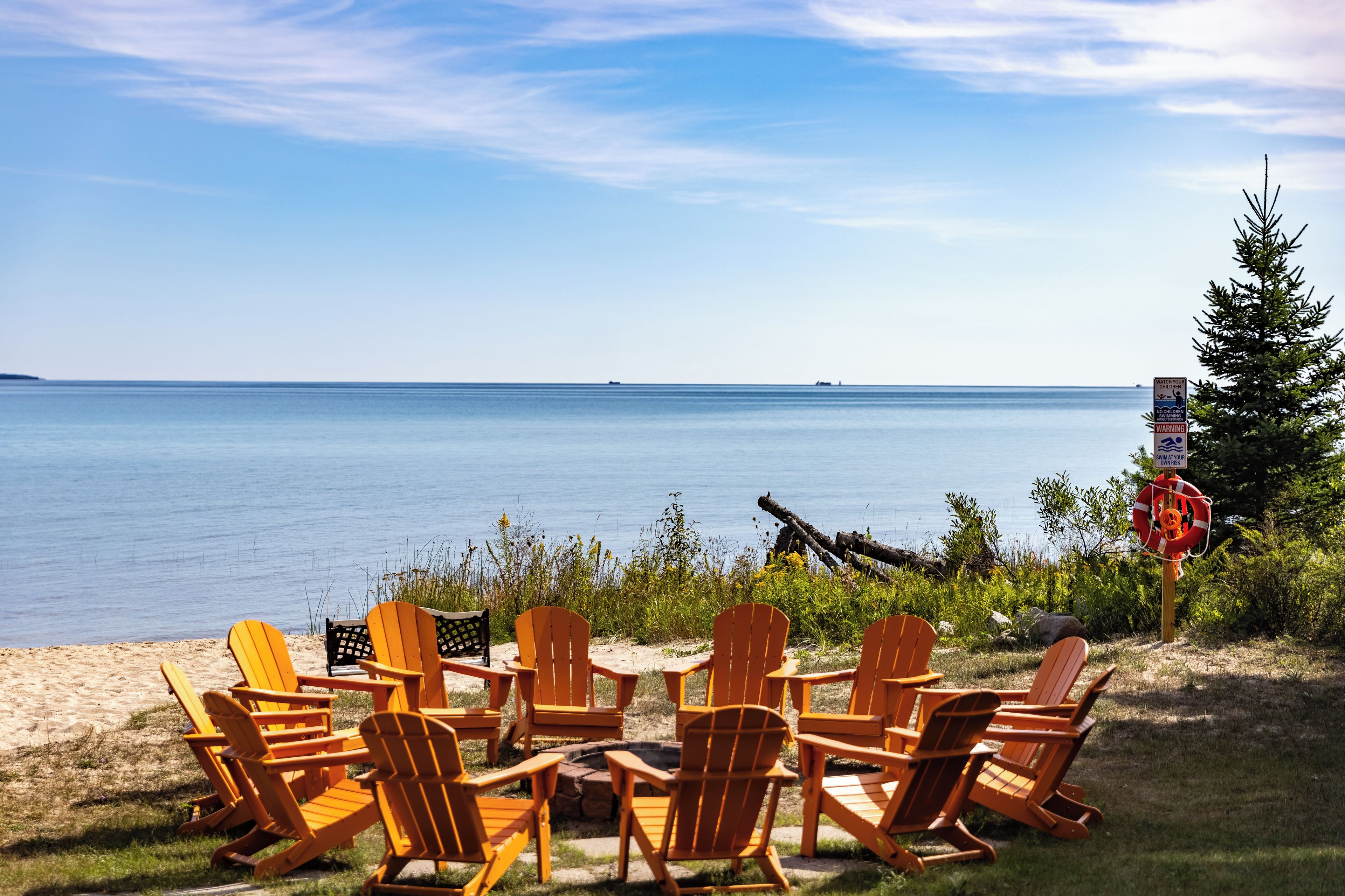 A beachside seating area with wooden chairs and a fire pit, overlooking a calm sea.