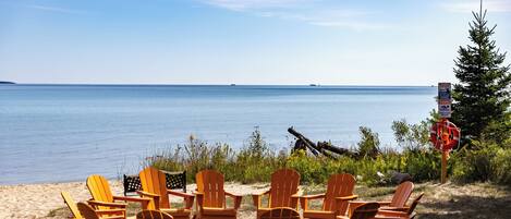 A beachside seating area with wooden chairs and a fire pit, overlooking a calm sea.