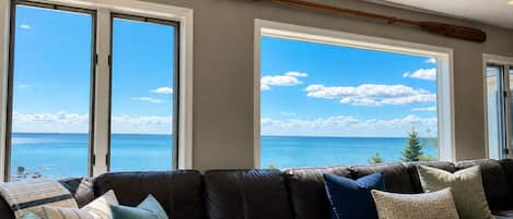 Un canapé en cuir agrémenté de plusieurs coussins, de grandes fenêtres offrant une vue sur la mer et une batte de baseball en bois accrochée au mur.