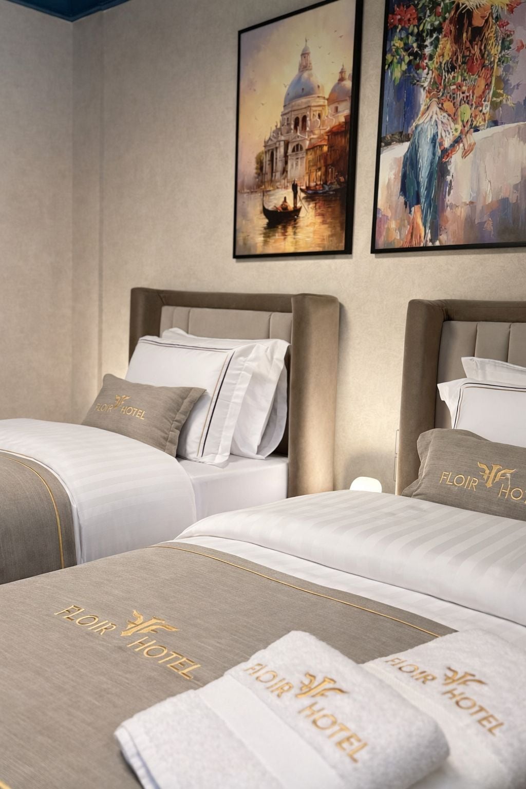 Habitación de hotel con dos camas, cada una con cabecero, ropa de cama blanca y almohadas con el logotipo del hotel. Hay dos obras de arte enmarcadas en las paredes, una que representa un edificio y otra un cuadro abstracto de colores.