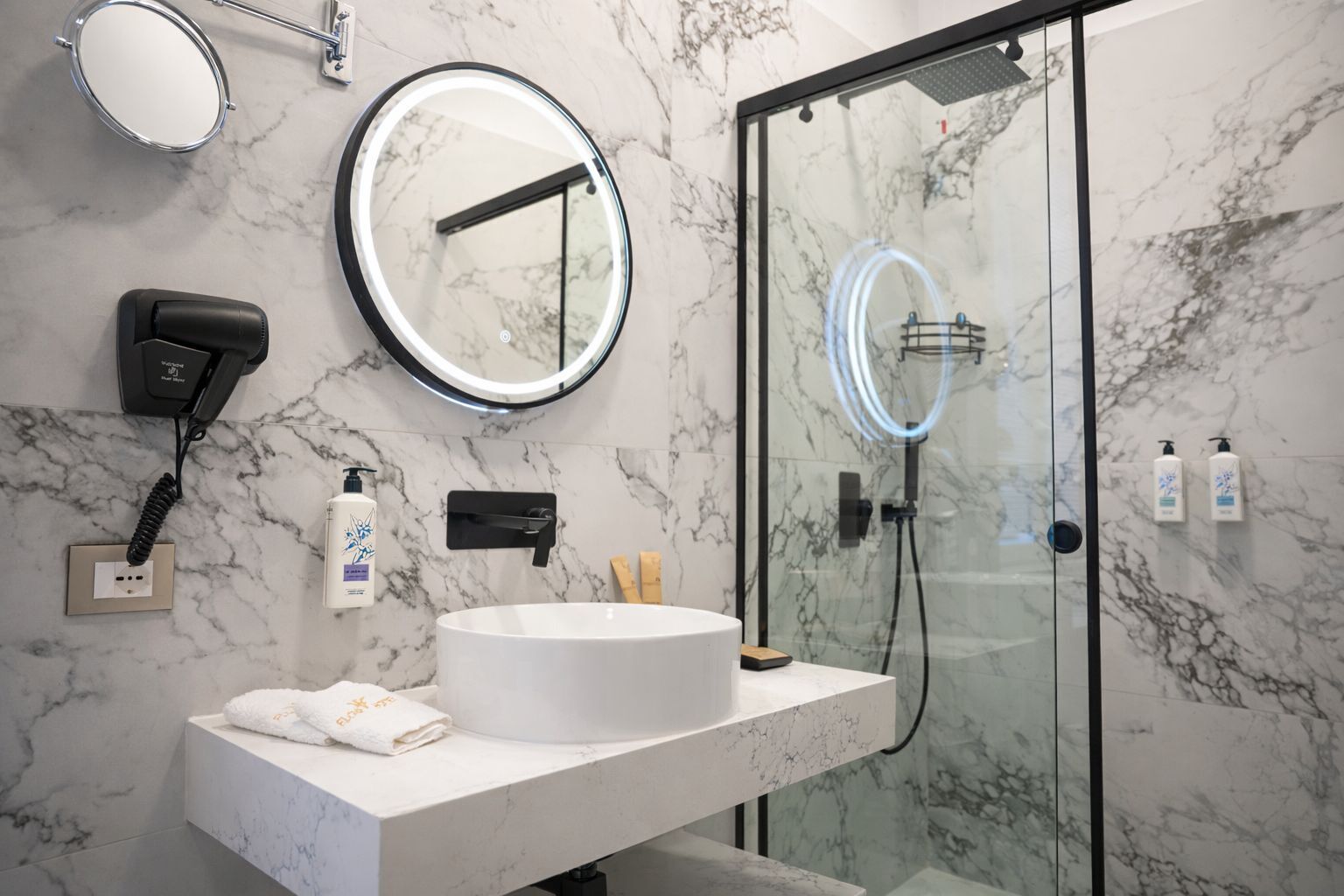 A modern bathroom with a marble sink, a round mirror, a hairdryer, and a glass-enclosed shower.