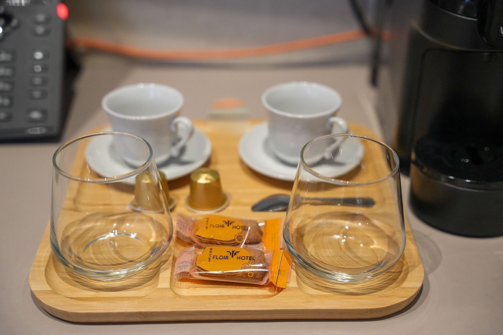 A tray with two clear glasses, two white cups, a small gold container, and a packet labeled 'FLOIR HOTEL.'