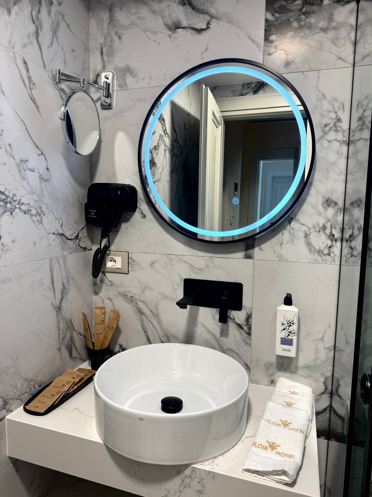 Uma casa de banho com uma pia branca, um espelho redondo com luz azul, um secador de cabelo e um porta-toalhas com uma toalha.