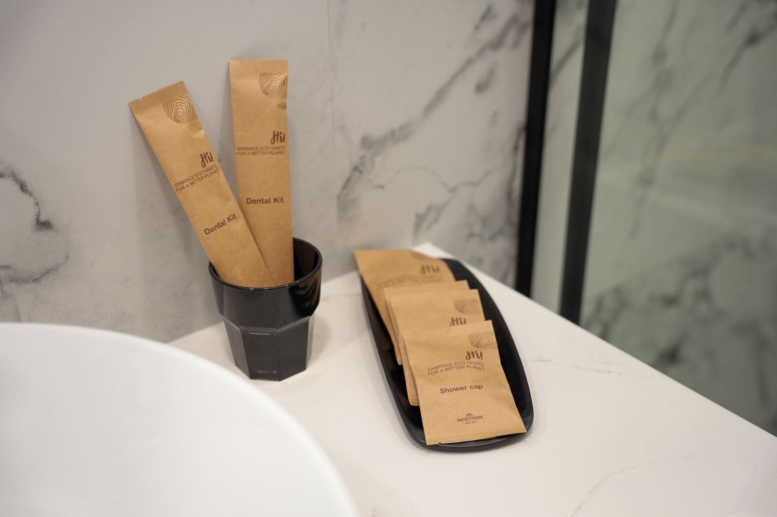 A bathroom with a marble sink, a black cup, and paper bags labeled 'Dental Kit' and 'shower cap.'