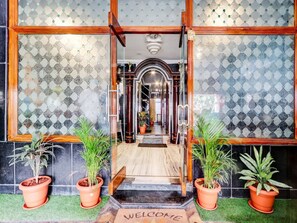 Entrada de un hotel con un felpudo que dice 'BIENVENIDOS', plantas en macetas y una pared con diseño.