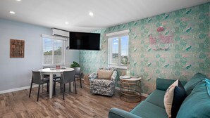 Een woonkamer met een blauwgroene bank, een eettafel met stoelen, een aan de muur bevestigde televisie en behang met een patroon.