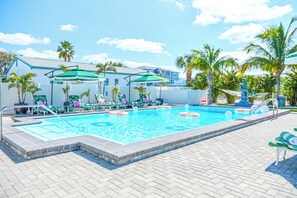Una piscina con sillones de descanso y sombrillas, rodeada de palmeras y un edificio con techo azul.