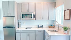 A modern kitchen with grey cabinets, a stainless steel refrigerator, a microwave, a toaster, and a sink. - Unit 104 King Suite 1 Bed/1Bath - Partial view - The Pearl Cabana Club (St Pete Beach)