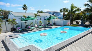 A swimming pool with lounge chairs and umbrellas, surrounded by palm trees and a building. - Unit 105 King Suite 1 Bed/1 Bath - The Pearl Cabana Club (St Pete Beach)