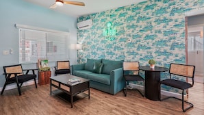 A living room with a teal sofa, black chairs, a coffee table, a ceiling fan, and a wall with a marine-themed wallpaper. - Unit 105 King Suite 1 Bed/1 Bath - The Pearl Cabana Club (St Pete Beach)