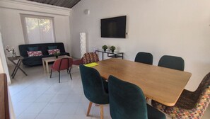 A dining area with a wooden table, green upholstered chairs, a sofa with floral cushions, and a TV mounted on the wall.
