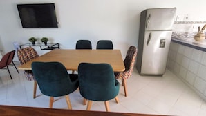 A dining area with a wooden table and chairs, a television mounted on the wall, and a refrigerator in the kitchen area.