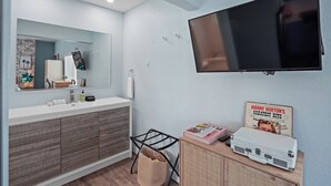 Une salle de bain moderne avec un grand miroir, un téléviseur à écran plat, un meuble-lavabo et une valise posée sur un meuble.