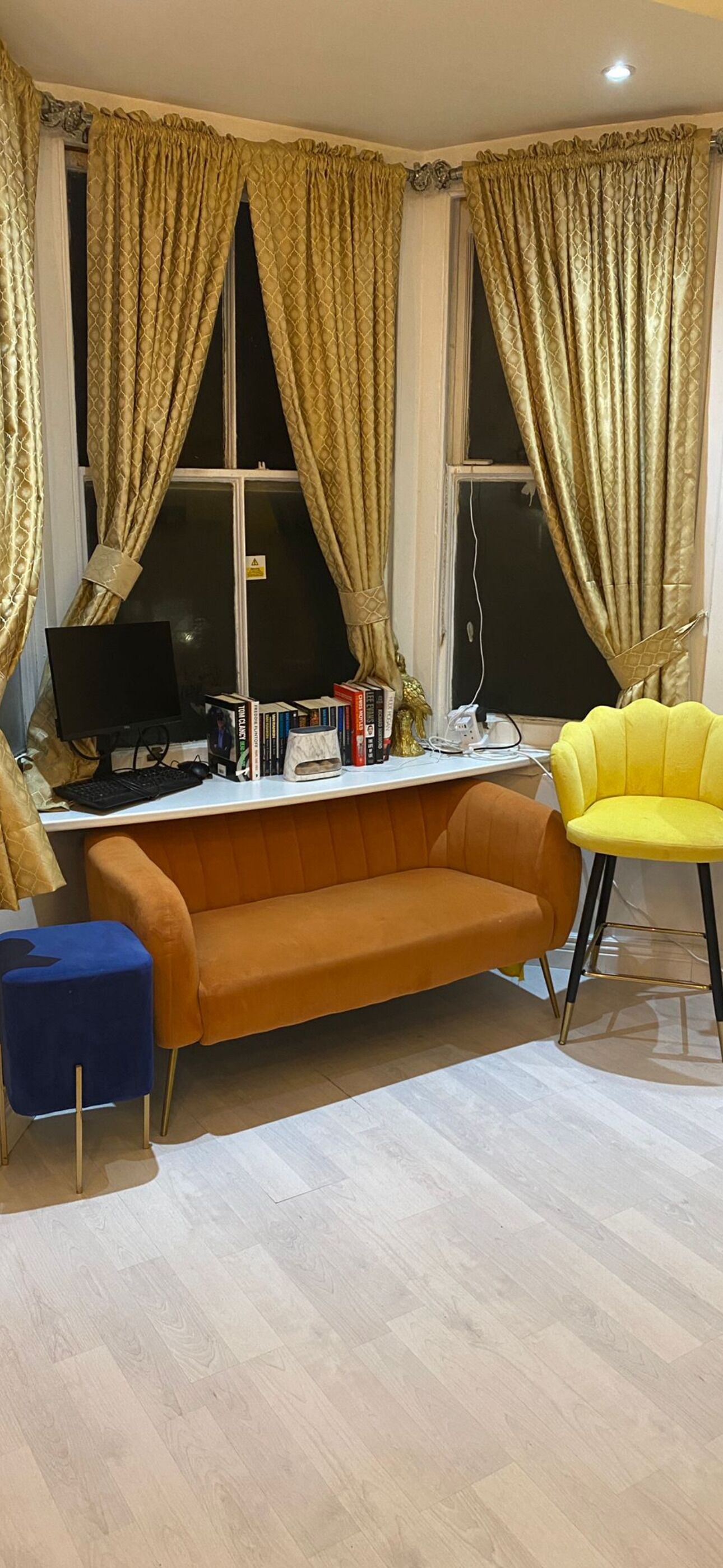 A room with a desk, computer, books, a yellow chair, an orange sofa, and large windows with curtains.