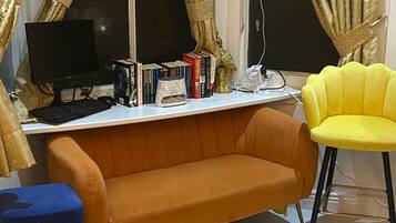 A room with a desk, computer, books, a yellow chair, an orange sofa, and large windows with curtains.