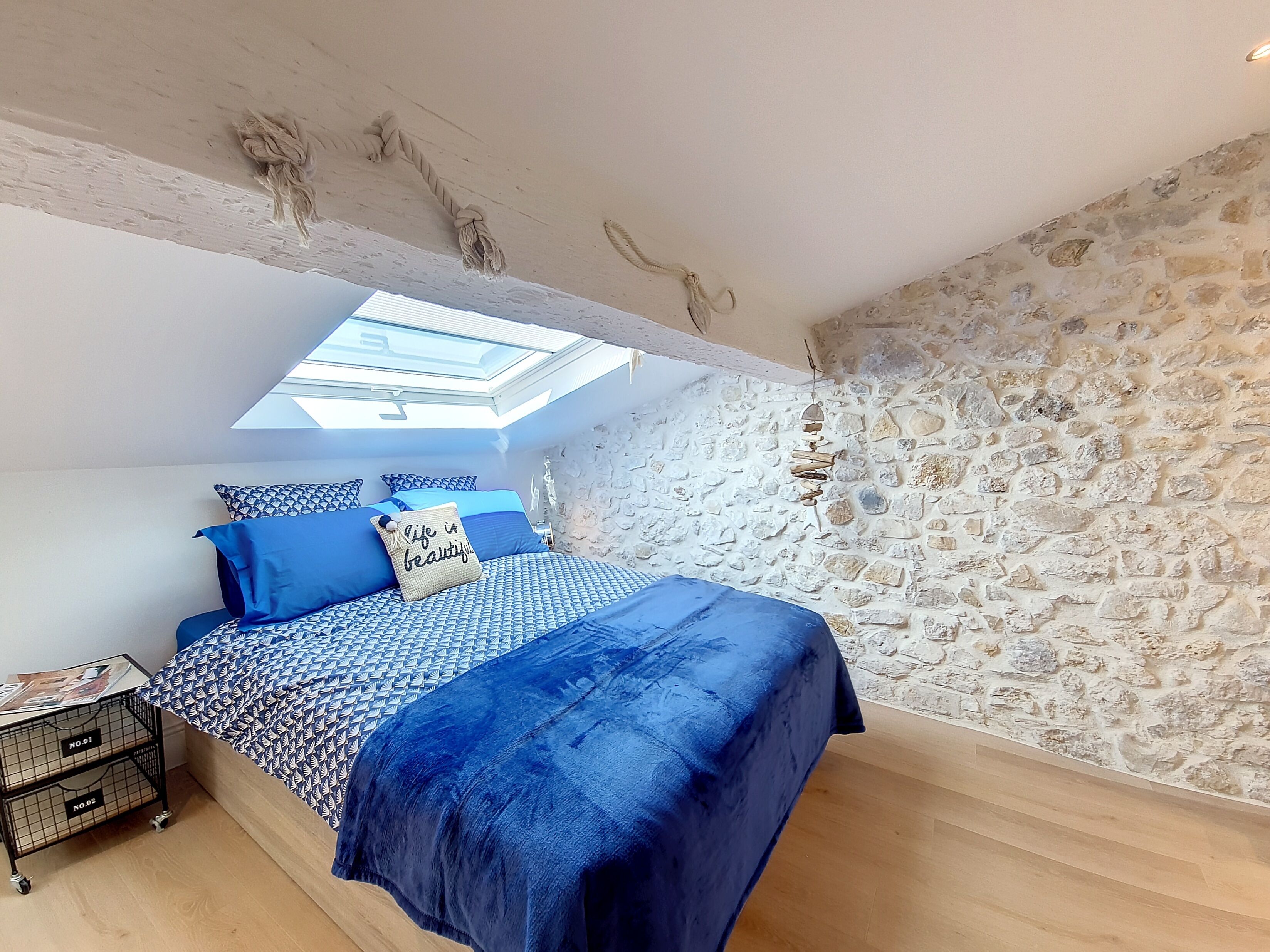 Un dormitorio con tragaluz, una pared de piedra, una cama con ropa de cama azul y una pequeña mesa con una lámpara.