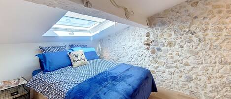Un dormitorio con tragaluz, una pared de piedra, una cama con ropa de cama azul y una pequeña mesa con una lámpara.