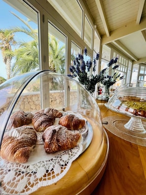 A breakfast buffet with pastries under a glass dome, a lace doily, and a view of palm trees through large windows.