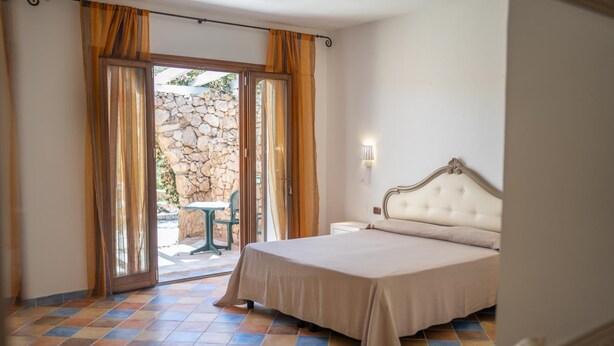 Une chambre avec un lit, une table de chevet, une lampe fixée au mur et une vue sur un espace extérieur accessible par une porte vitrée, cet espace étant agrémenté d’un mur en pierre et d’une table.