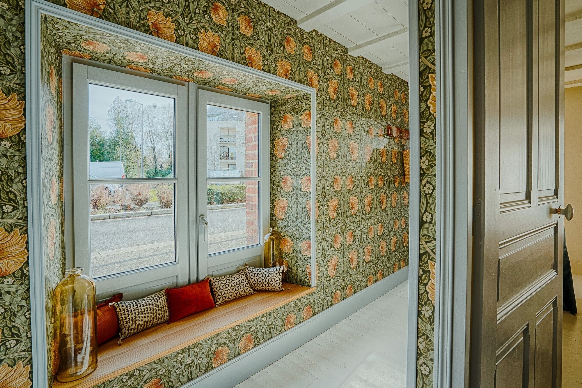 Une pièce avec une fenêtre, un banc agrémenté de coussins et un papier peint à motifs floraux.