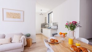 Een moderne woonkamer met een bank, een eettafel met croissants en sap, en een keuken op de achtergrond.