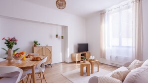 Een moderne woonkamer met een houten eettafel, witte stoelen, een bank, een televisie en een raam met dunne gordijnen.
